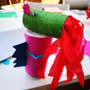 Slika prikazuje zelenega in roza zmaja, ki bruha ogenj, narejenega iz tulcev za straniščni papir in krep papirja.