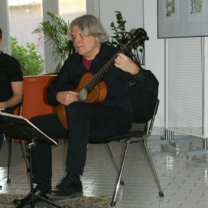 Slika prikazuje sedečega glasbenika s kitaro v rokah.